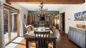 A dining area with a wooden table, chairs, and a chandelier. There is a kitchen in the background with a TV and cabinets. - Snow Haus | Hot Tub & Bar | 10 Min to Mount Snow! (Wardsboro)