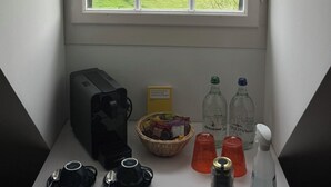 A room with a large window overlooking a green landscape and water, a small table with a basket of items, and a coffee maker. - Hotel St. Petersinsel (Twann)