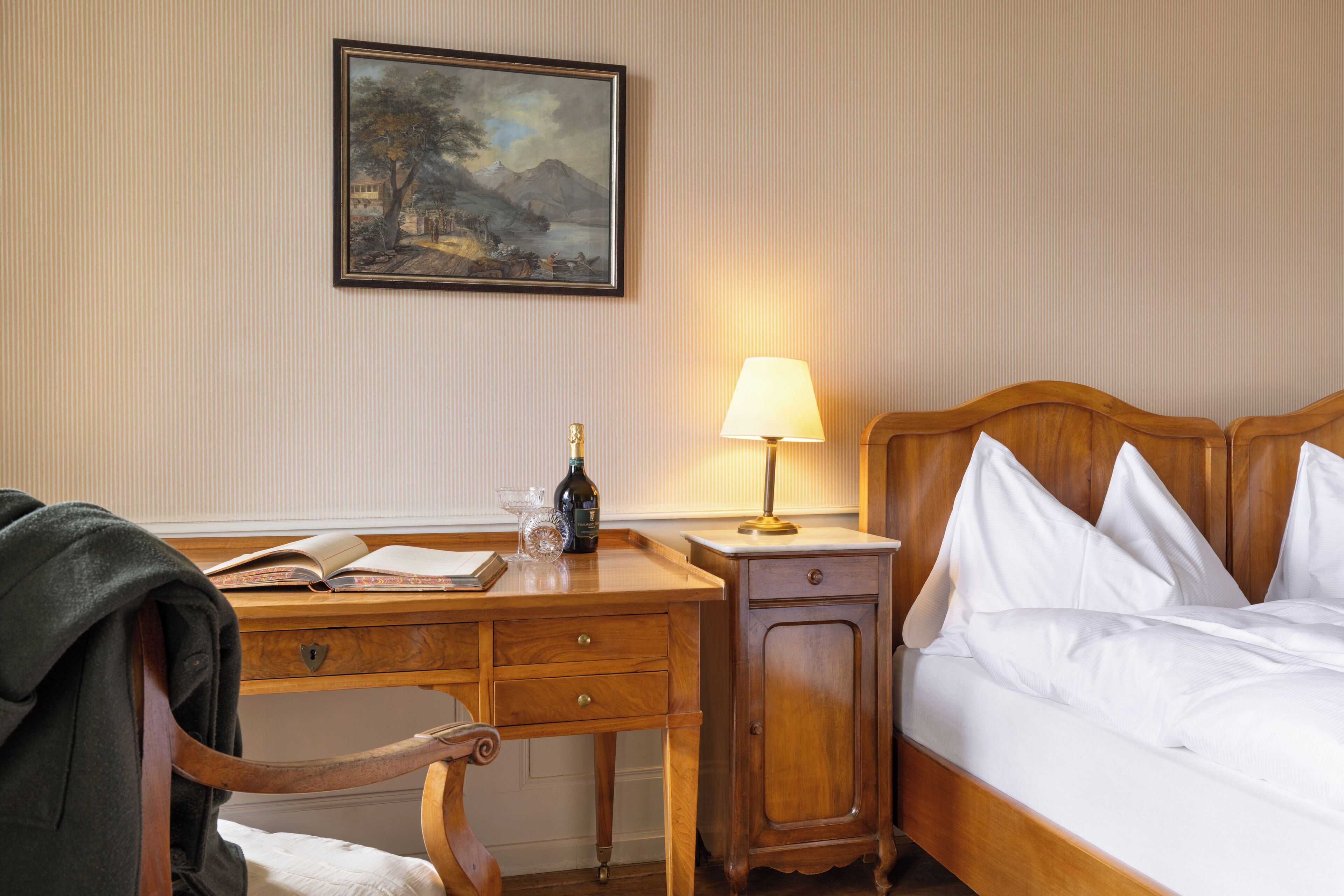 A hotel room with a wooden desk, a bed with white linens, a nightstand with a lamp, and a framed painting on the wall.