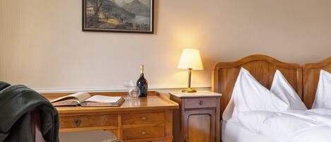 A hotel room with a wooden desk, a bed with white linens, a nightstand with a lamp, and a framed painting on the wall.