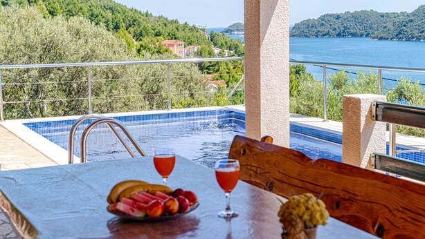 Une table placée au bord de la piscine, avec des fruits et des boissons, une vue sur un lac et un paysage montagneux.