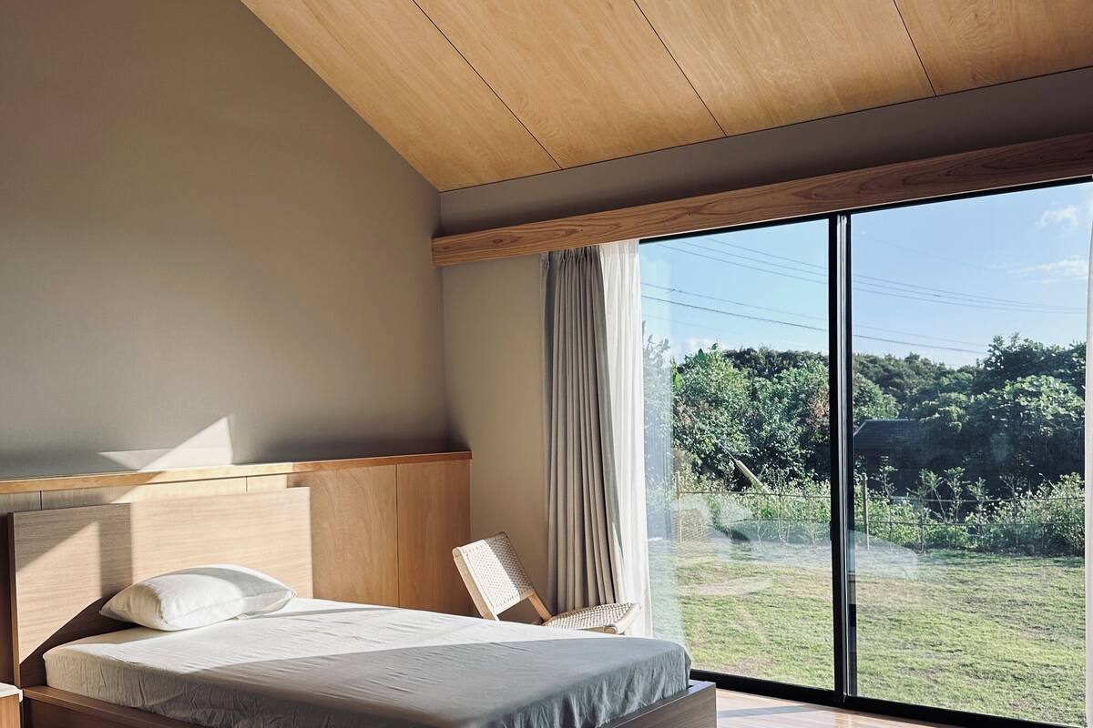 Un dormitorio moderno con una cama grande, techo de madera y vista a un paisaje verde a través de amplios ventanales.