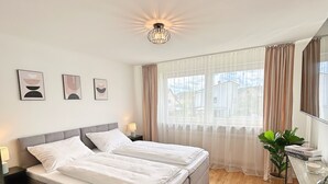 Ein Schlafzimmer mit Doppelbett, Nachttisch mit Lampe, Fenster mit Vorhängen und einer Pflanze.