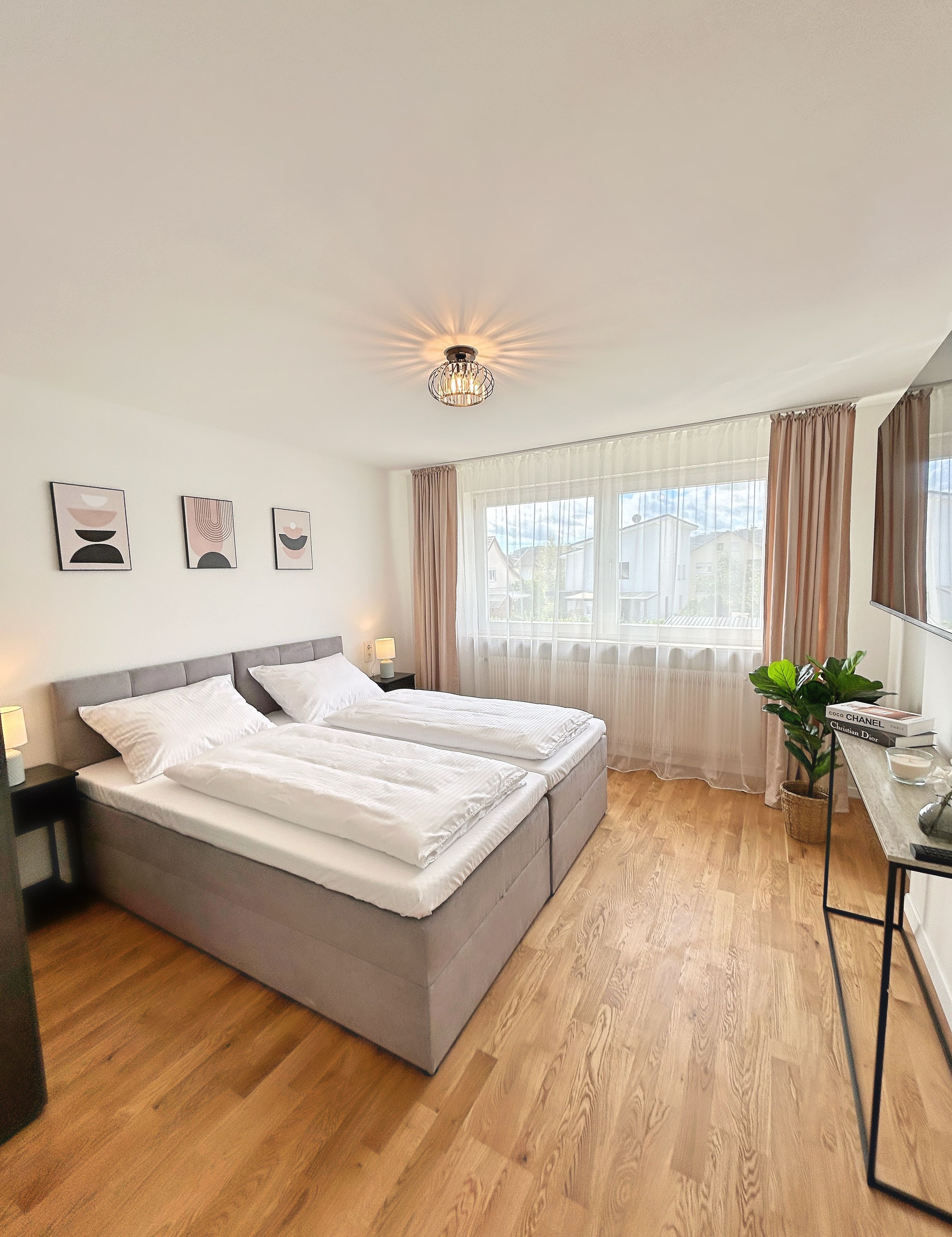 Ein Schlafzimmer mit Doppelbett, Nachttisch mit Lampe, Fenster mit Vorhängen und einer Pflanze.