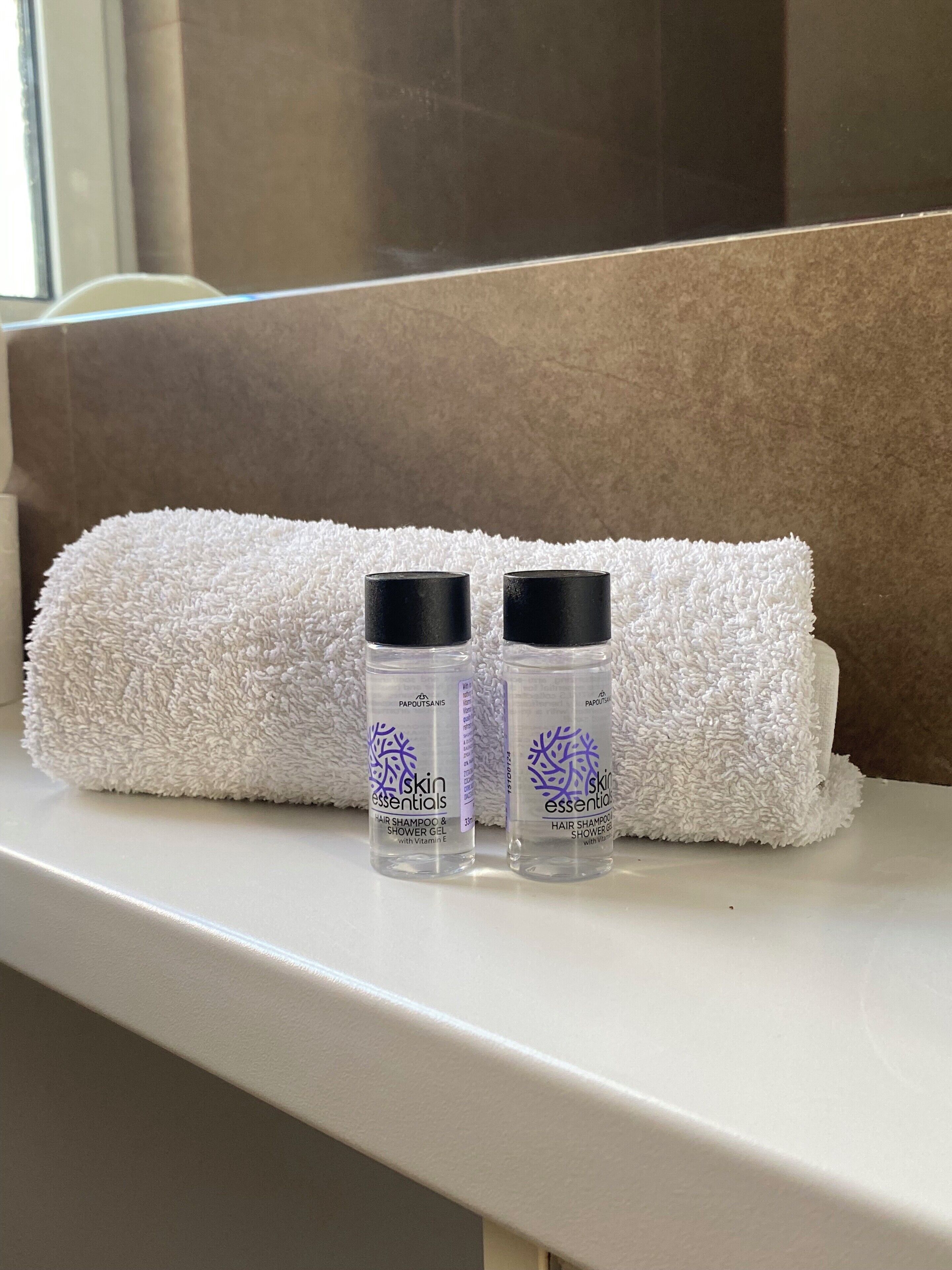 A bathroom with a white towel rolled up and two bottles of skin essentials hair shampoo and shower gel on a white shelf.
