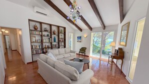 Ein geräumiges Wohnzimmer mit einer großen Couch, einem Couchtisch und einem Bücherregal voller Bücher und dekorativer Gegenstände. Ein Kronleuchter hängt von der Decke, und große Fenster sorgen für natürliches Licht und Blick ins Freie.