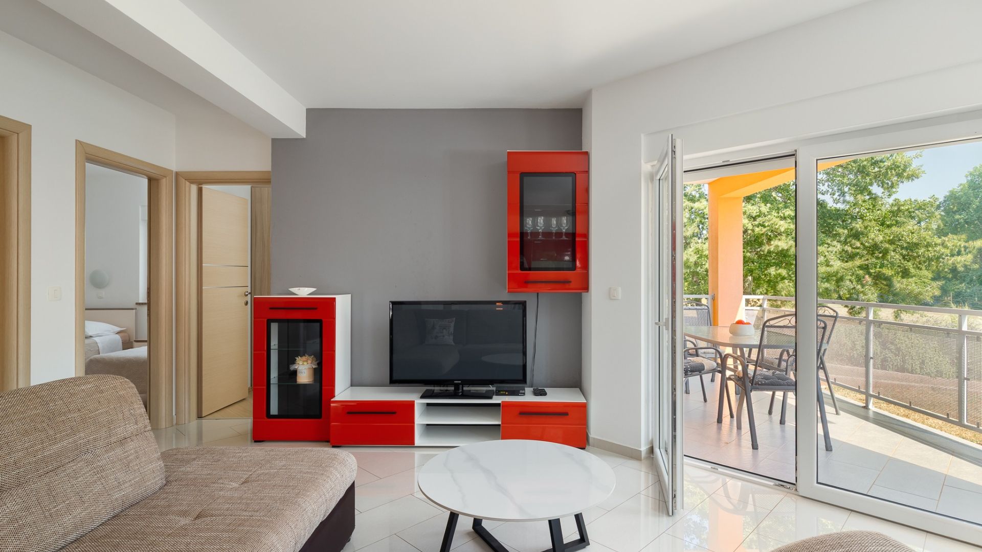 Un salon moderne doté d’un téléviseur à écran plat, d’un meuble rouge et d’un balcon aménagé avec du mobilier d’extérieur.