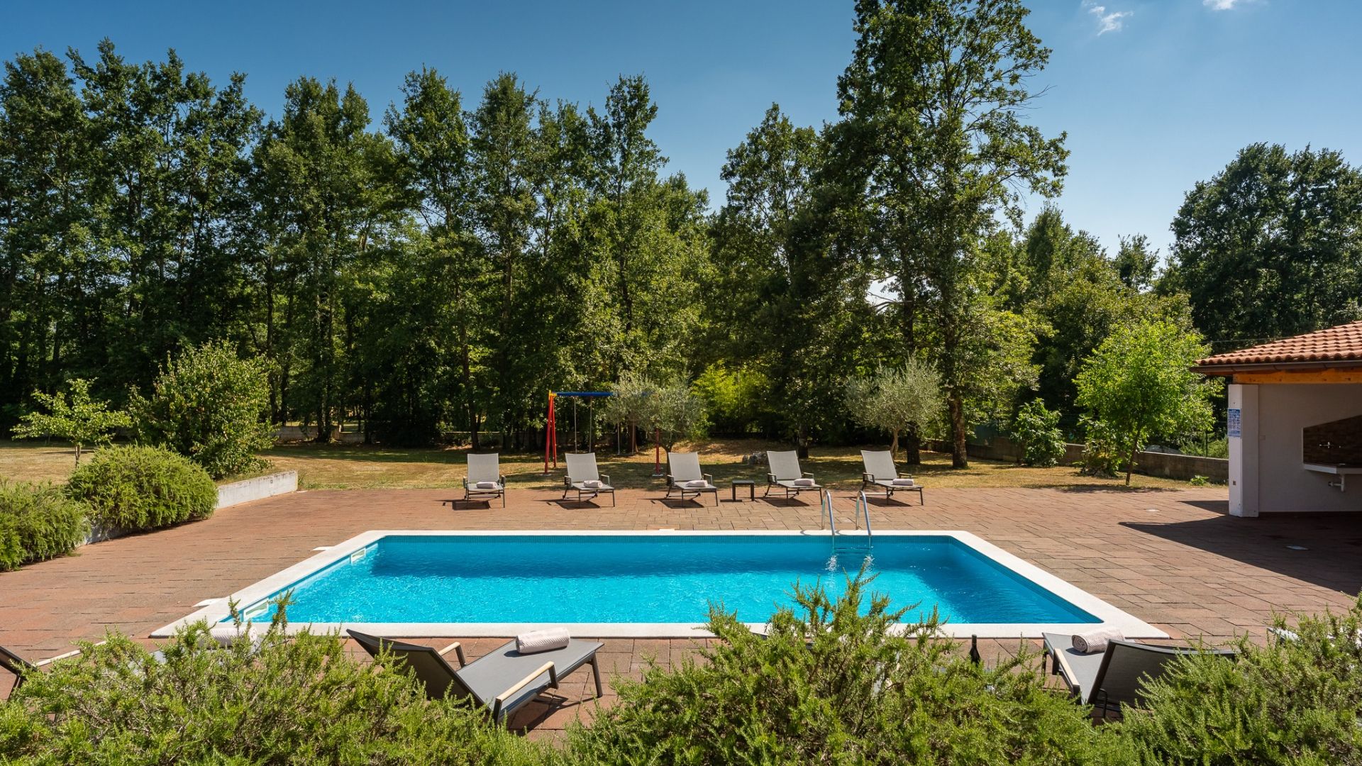 Une piscine rectangulaire avec une terrasse carrelée, entourée d’arbres et dotée de plusieurs chaises longues.