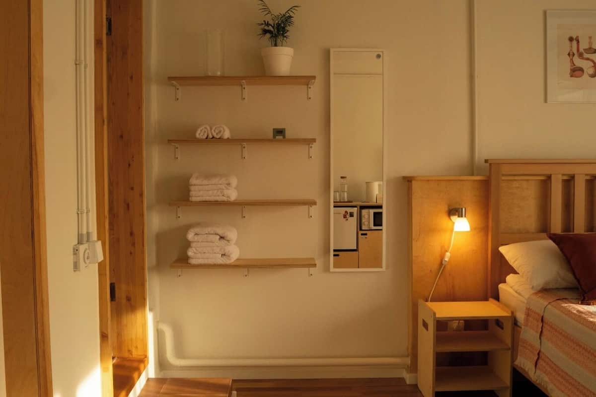 Makuuhuoneessa on sänky, yöpöytä, lamppu, pyyhkeillä täytetyt hyllyt, ruukkukasvi, peili ja jääkaappi.