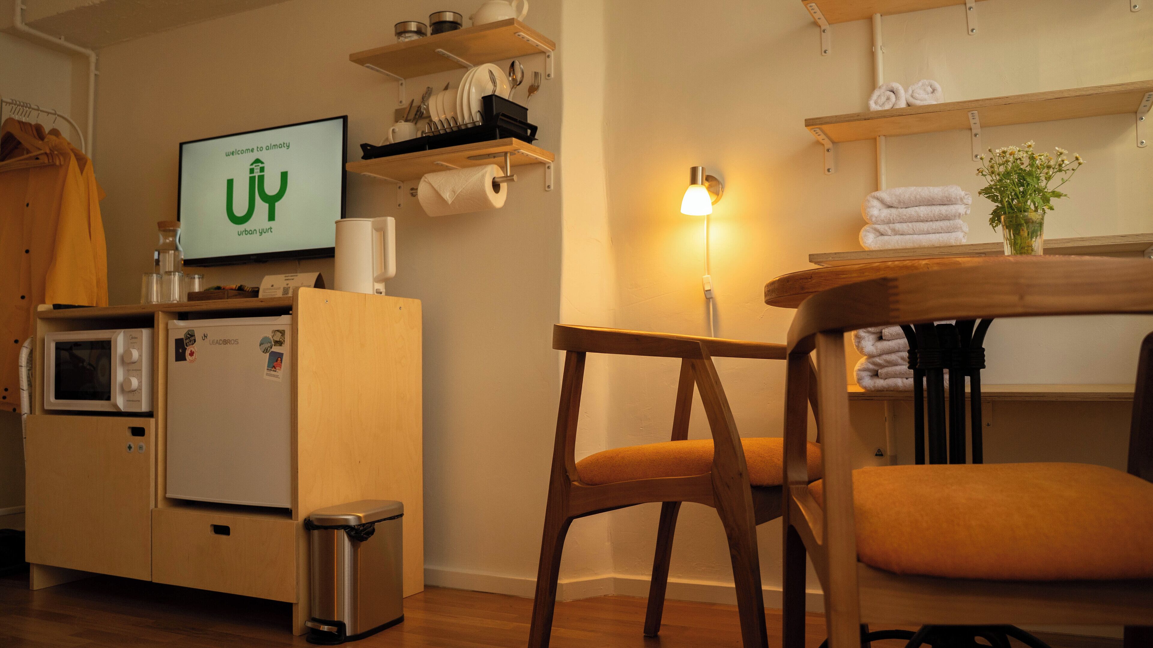 Una kitchenette compacta con microondas, un refrigerador pequeño y un cesto de basura. Hay una silla de madera y una mesa con una lámpara. Un televisor muestra el logotipo de 'almaty'.