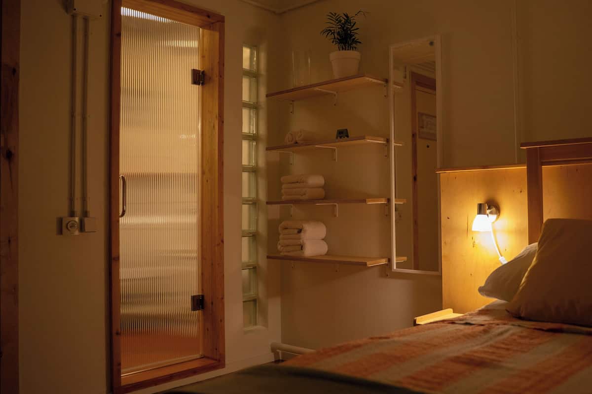 Hotellihuone, jossa on sänky, yöpöytälamppu, hylly pyyhkeille ja puinen ovi, jossa on sälekaihtimet.