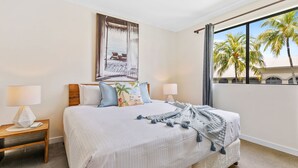 Une chambre avec un grand lit, deux tables de chevet avec des lampes, une fenêtre donnant sur des palmiers et un tableau représentant une scène de plage accroché au mur.