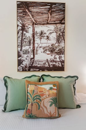 A framed tropical scene with palm trees and a pathway, a green pillow with palm tree design, and a white bed with a striped pillow. - Palm Cove Studio Pools Spa Bar (Cairns)