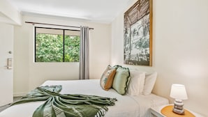 A neatly made bed with a green throw blanket, a bedside table with a lamp, and a window with curtains. - Palm Cove Studio Pools Spa Bar (Cairns)