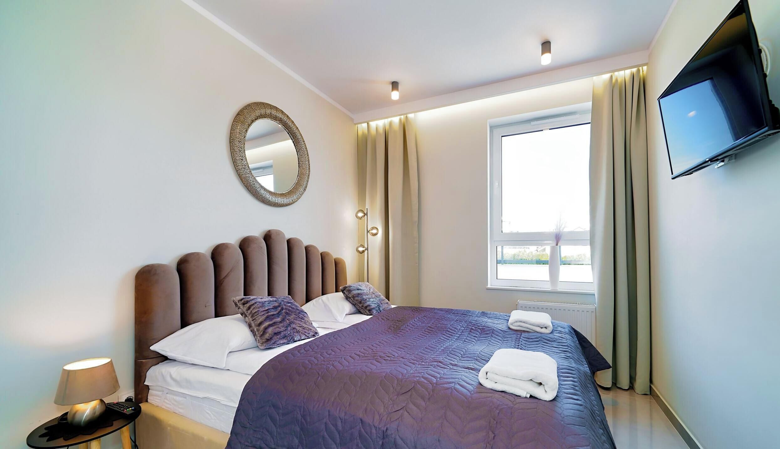 A modern hotel room with a large bed, a flat-screen TV mounted on the wall, a bedside table with a lamp, and a window with curtains.