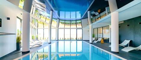 An indoor swimming pool with clear glass walls, white lounge chairs, and a modern architectural design.