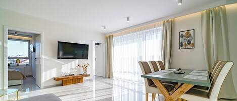 A modern living room with a dining area, a flat-screen TV mounted on the wall, and a large window with sheer curtains.