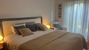 Ein ordentlich bezogenes Bett mit gemustertem Überwurf und Kissen, eine Nachttischlampe und ein Sessel mit Sitzkissen.