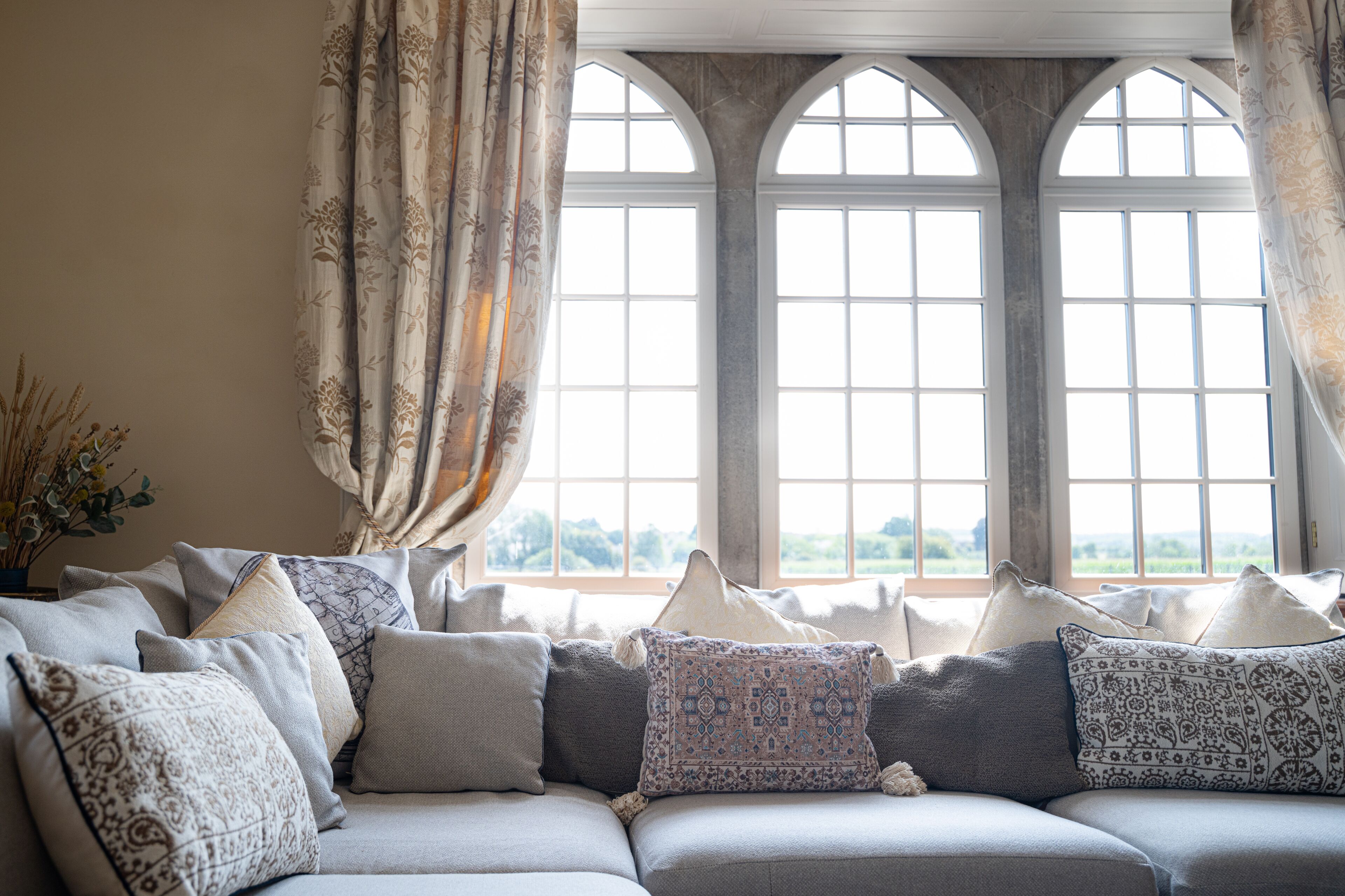 A cozy living room with a sectional sofa adorned with patterned cushions, large arched windows, and a view of greenery outside.