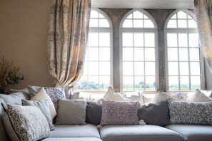 A cozy living room with a sectional sofa adorned with patterned cushions, large arched windows, and a view of greenery outside. - Manor Farmhouse, Holt, Wiltshire (Near Bath) (Holt)