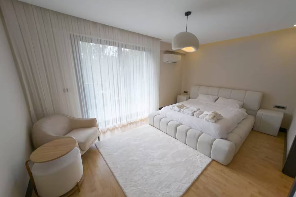 Une chambre moderne avec un grand lit, une chaise, une table ronde et une grande fenêtre ornée de rideaux légers.