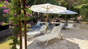Un patio exterior con sillones de descanso y un gran paraguas, rodeado de follaje y suelo de piedra.