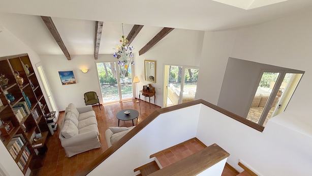 Amplia sala de estar con techo alto, vigas de madera, una escalera y ventanas grandes que ofrecen vistas al exterior.