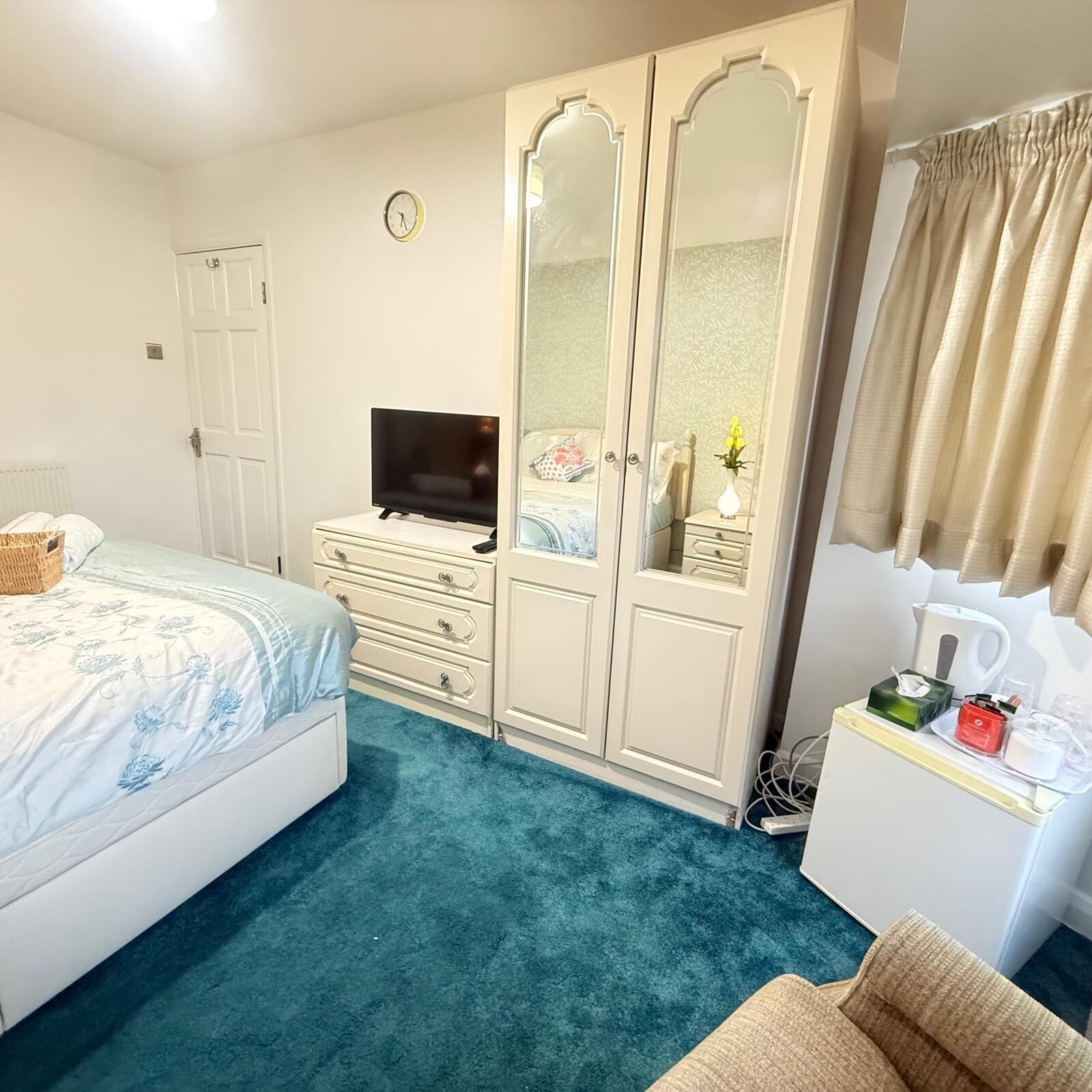 Ein Schlafzimmer mit Bett, Fernseher, Kleiderschrank, Kommode und Nachttisch mit Lampe und Blumen.