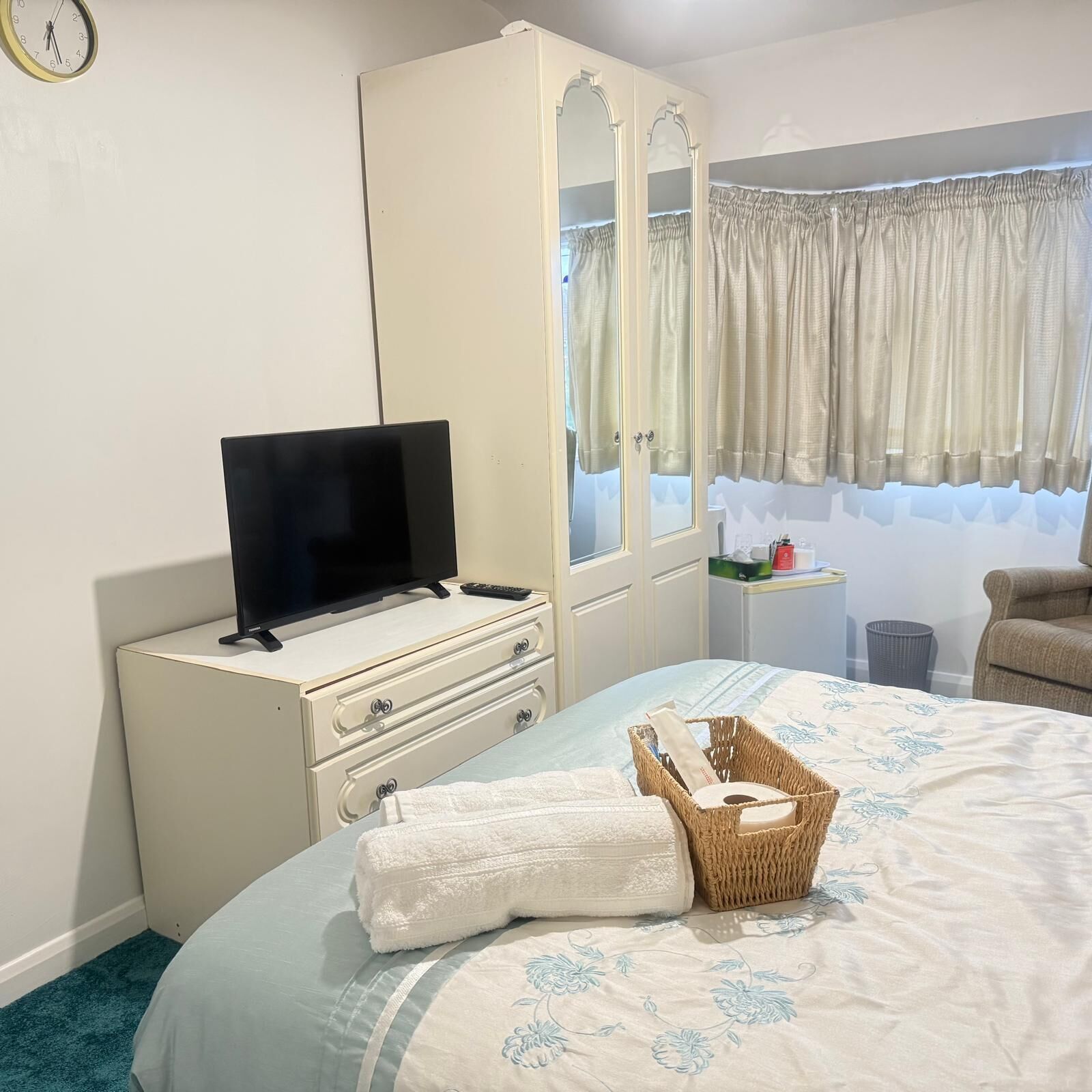A bedroom with a bed, a television on a dresser, a mirror, a window with curtains, and a wicker basket.