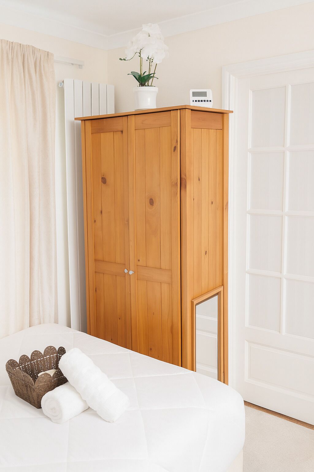 Un dormitorio con un armario de madera, una cama, una almohada blanca, una toalla blanca, una pequeña orquídea en maceta y un reloj digital en la pared.