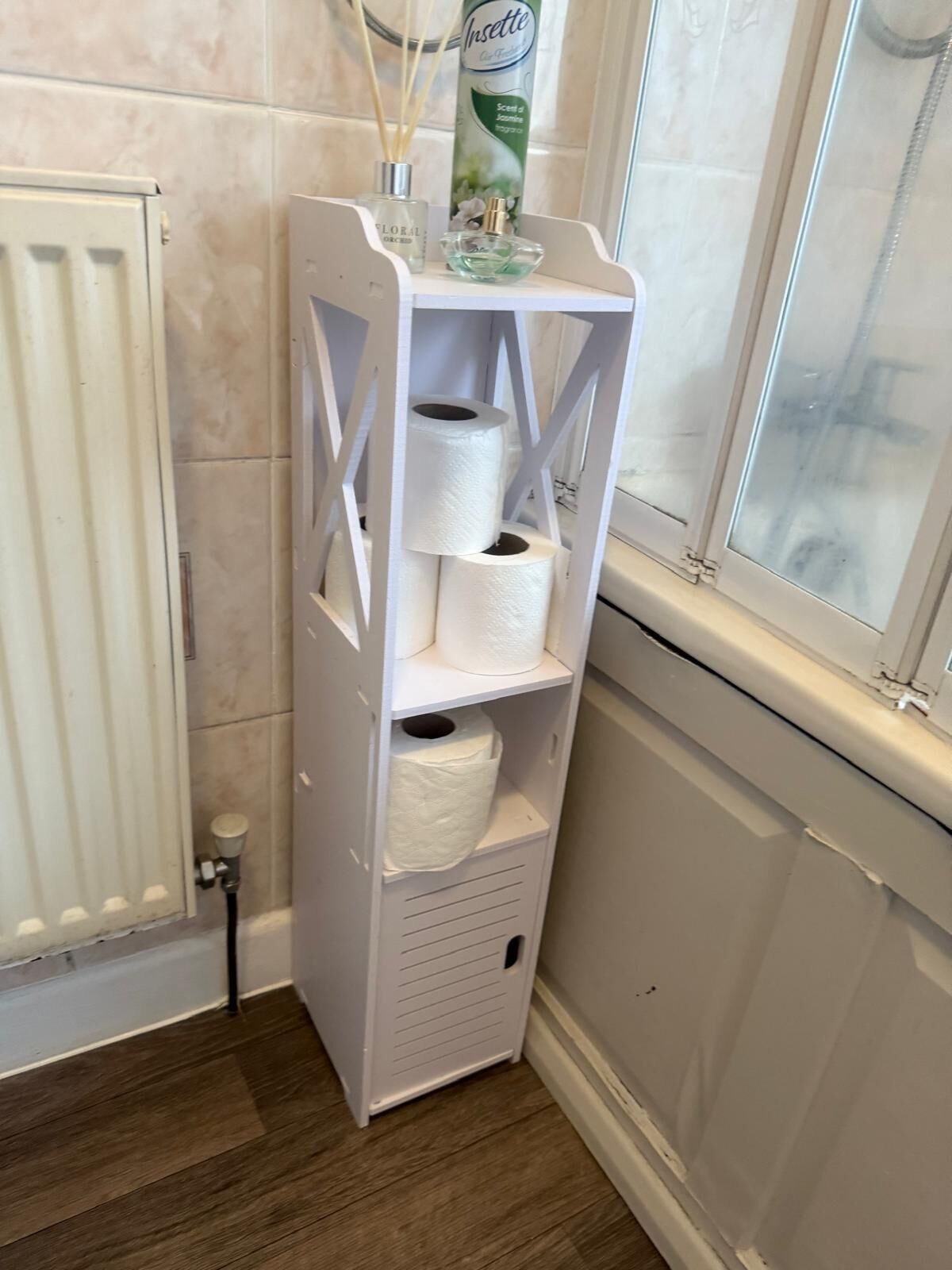 Un módulo de almacenamiento blanco con papel higiénico y un spray corporal Inselette en un baño con ventana y radiador.