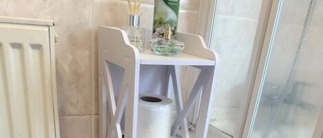 Un módulo de almacenamiento blanco con papel higiénico y un spray corporal Inselette en un baño con ventana y radiador.