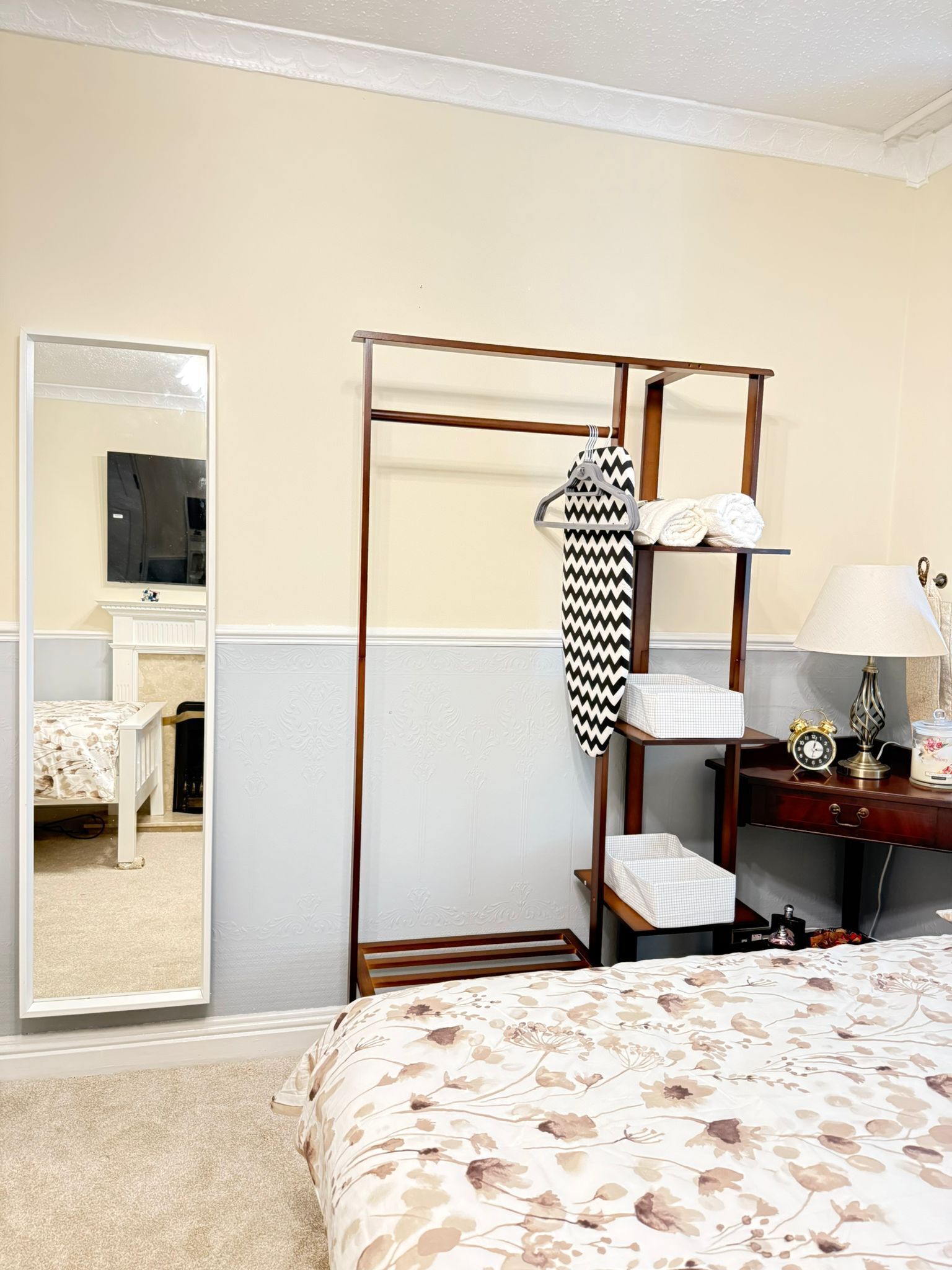 Un dormitorio con una cama, un armario de madera, un espejo, una mesita de noche con una lámpara y un reloj.