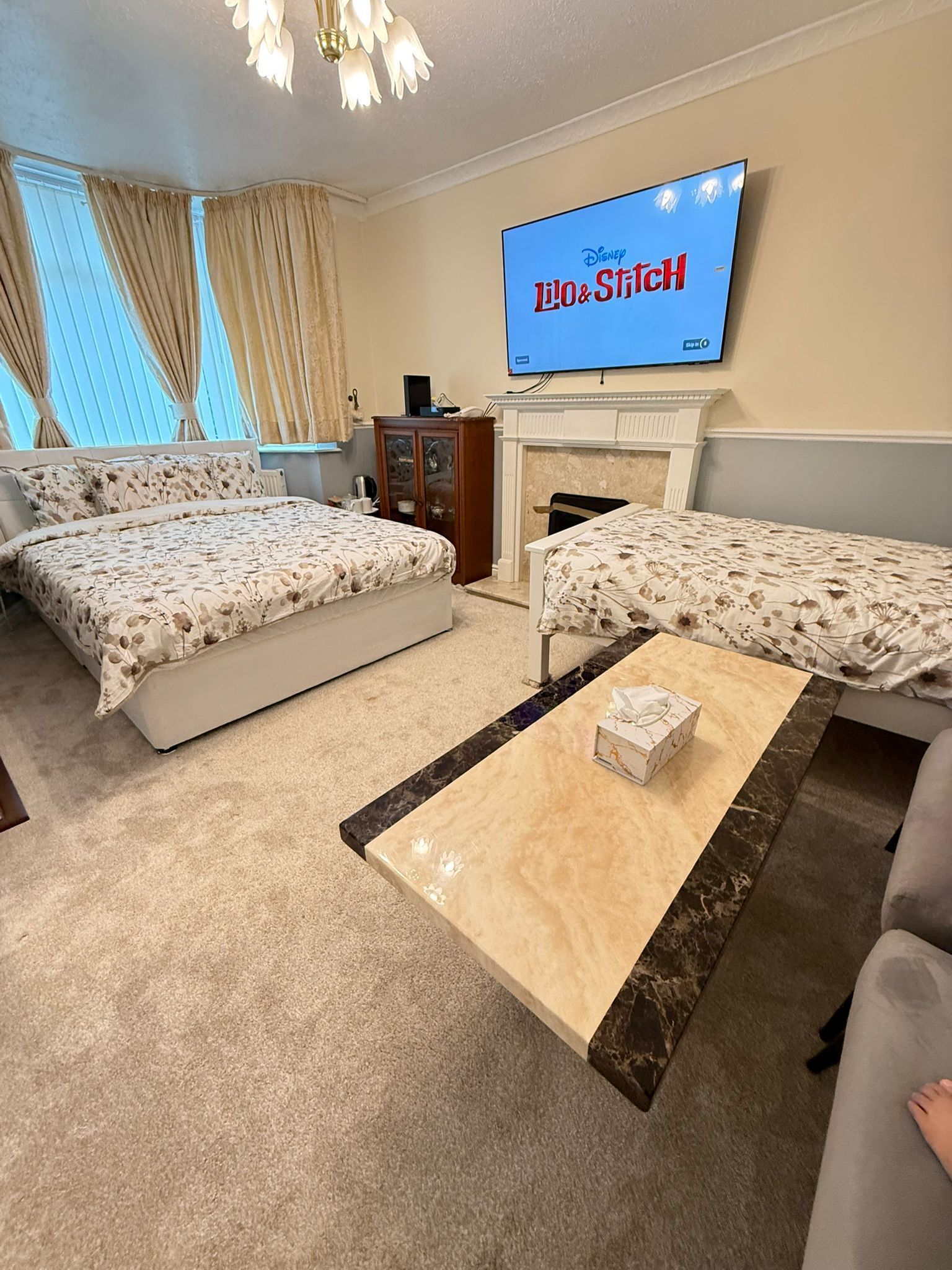 A hotel room with two beds, a TV displaying 'Disney Stitch', a fireplace, and a coffee table with a box.