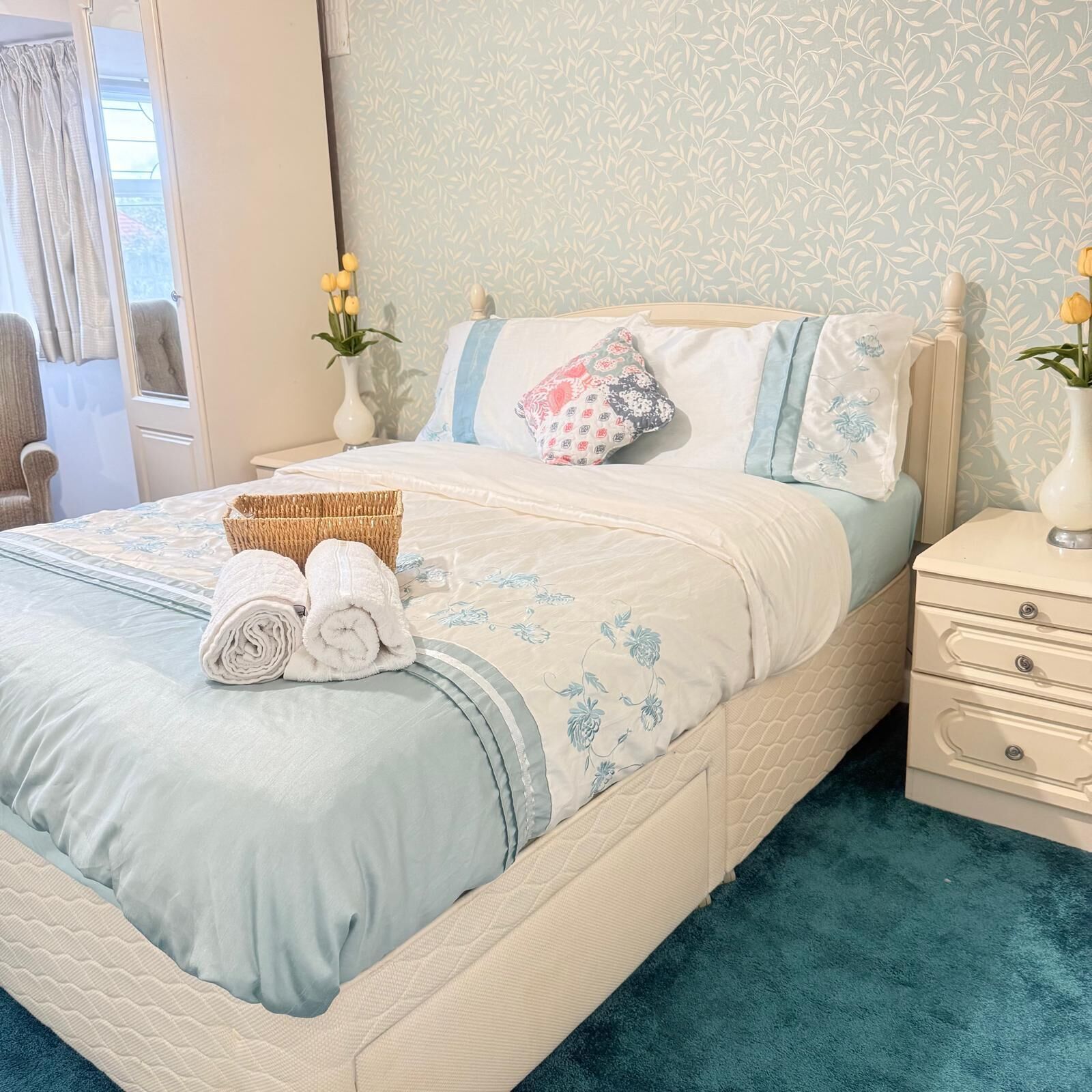 A neatly made bed with a blue and white floral bedspread, a bedside table with a vase of flowers, and a window with curtains.