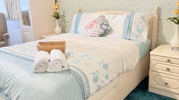 A neatly made bed with a blue and white floral bedspread, a bedside table with a vase of flowers, and a window with curtains.