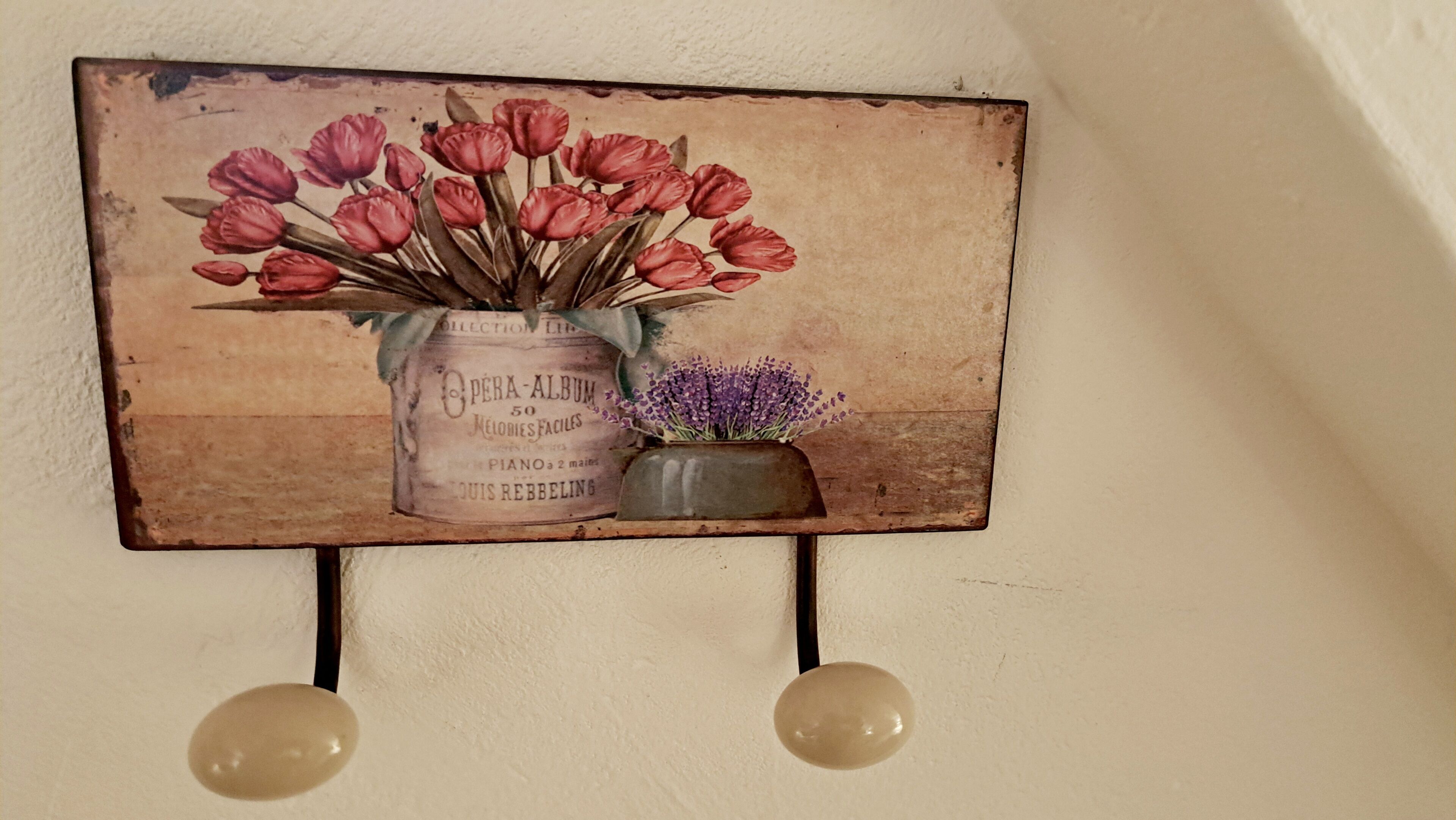 A vintage-style wall decoration with a floral arrangement in a tin can labeled "OPERA-ALBUM" and a small vase with purple flowers.