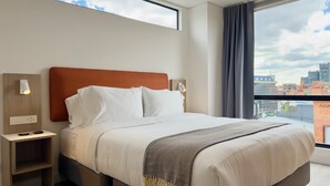Una habitación de hotel moderna con una cama grande, una mesita de noche, una lámpara de pared y vistas a la ciudad a través de un ventanal.