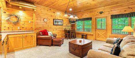 A cozy cabin interior with wooden walls, a fireplace, a dining area, and a living room with a sofa and a coffee table.