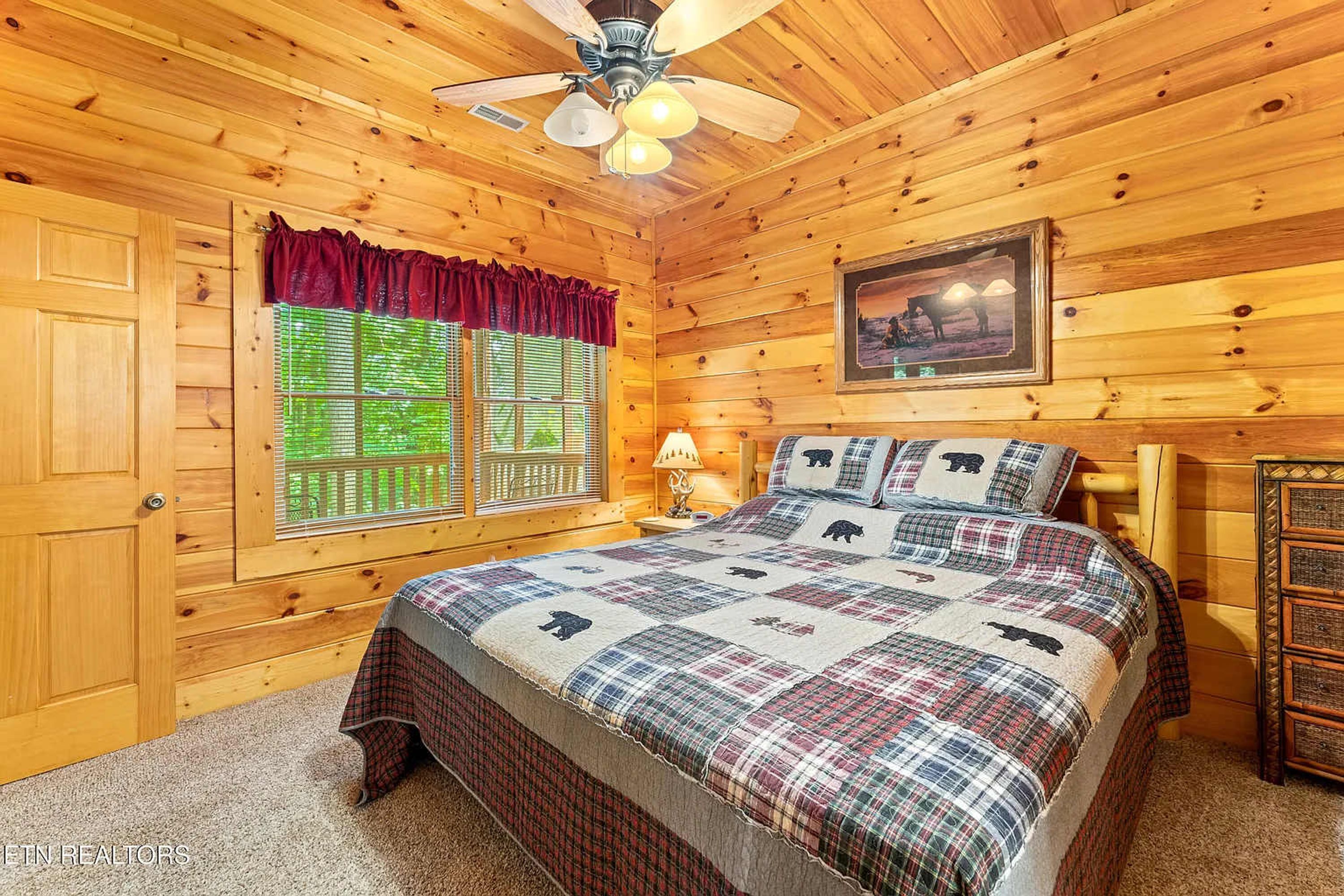 A cozy wooden cabin bedroom with a bed, a ceiling fan, a window with shutters, and a framed picture on the wall.