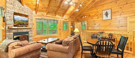 A cozy cabin interior with a stone fireplace, wooden walls, a ceiling fan, and a dining area with a table and chairs.