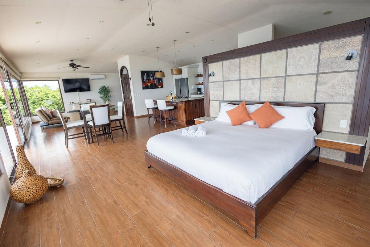 A spacious bedroom with a large bed, a dining area, and a kitchen in the background.