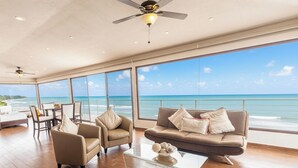 Una habitación amplia con un ventanal grande con vistas al océano, un ventilador de techo y una mesa de centro de cristal.