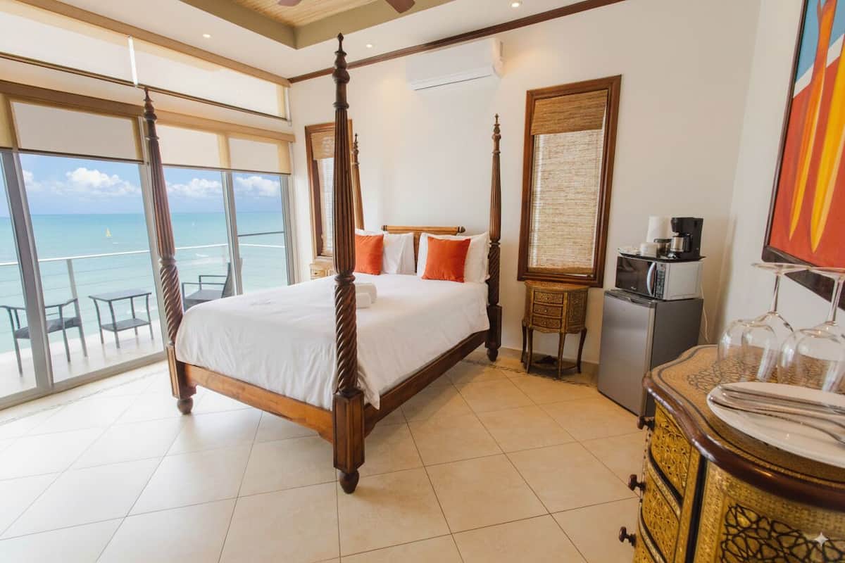 A four-poster bed with white linens, a small bedside table with a coffee maker, and a view of the ocean through large windows.