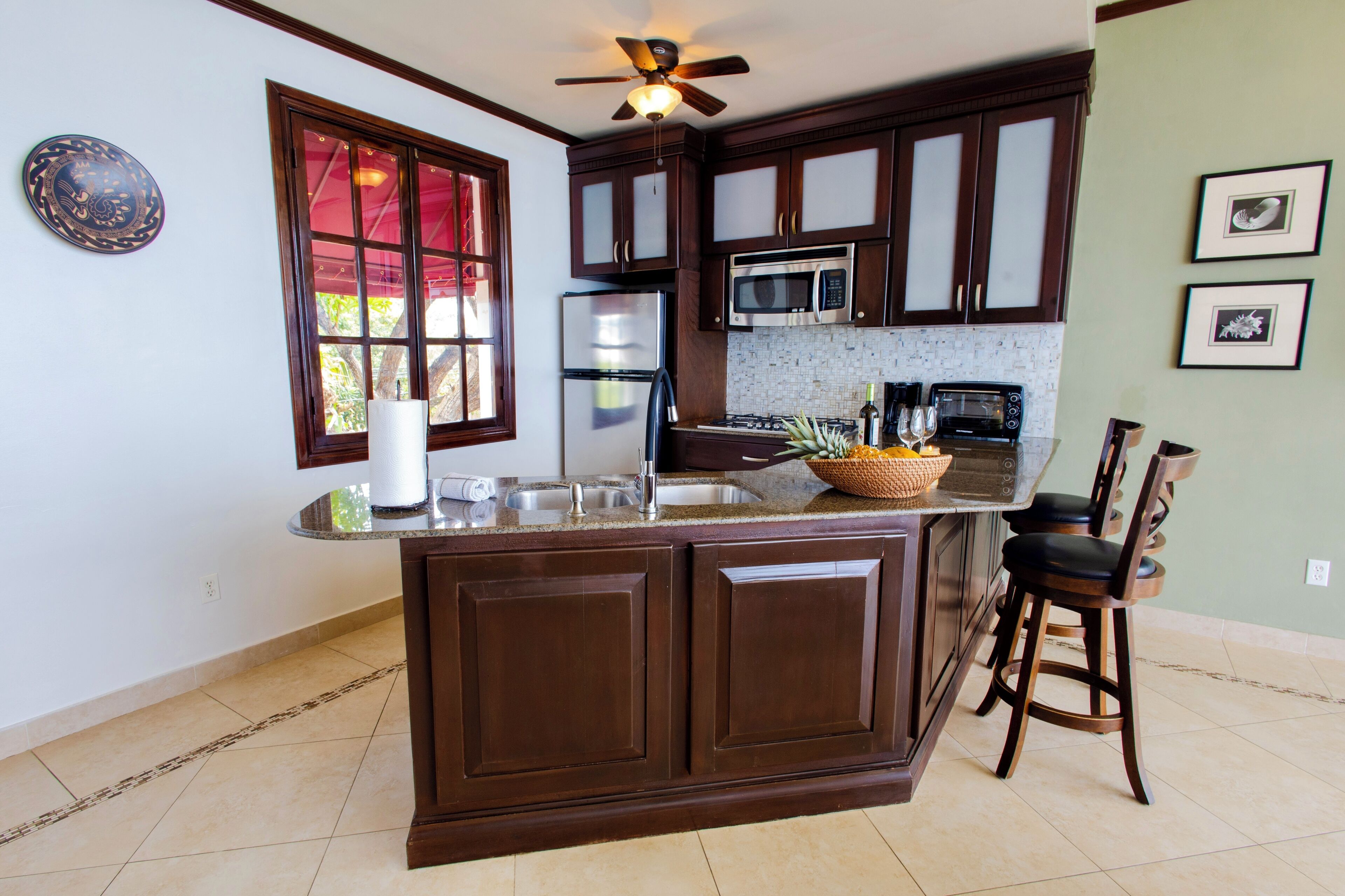 A kitchen with dark wooden cabinets, a central island, a microwave, a refrigerator, and a ceiling fan.
