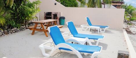 Un área de descanso frente a la playa con sillones blancos y azules, una mesa de madera y una parrilla.