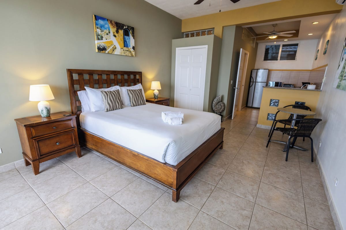 A bedroom with a wooden bed, two bedside tables with lamps, a white door, and a kitchenette in the background.