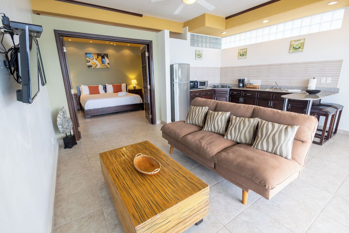 A modern living room with a sofa, coffee table, and a kitchen area visible through an open doorway.