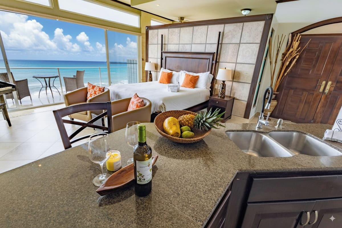 A hotel room with a bed, a dining area with a table and chairs, a kitchen with a sink and countertop, and a view of the ocean.