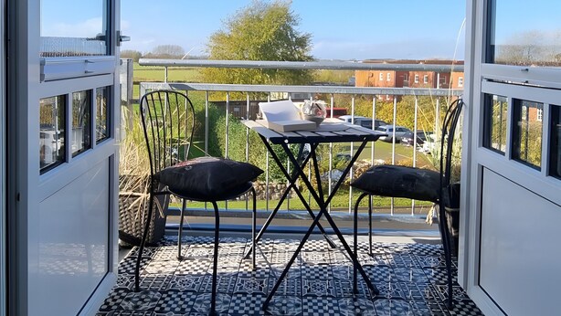 A balcony with a checkered floor, two chairs, a small table, and a view of a building and trees. - Apartment 'Inselstern' with Balcony and Wi-Fi (Pellworm)